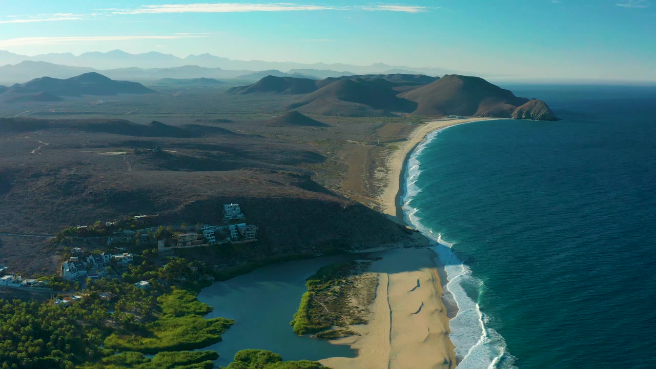 A high aerial frame in Todos Santos, Baja California Sur, holds the lagoon and vegetation at the lower edge, with a curving sandy shore, white surf lines, dry land, hills, and no visible people.