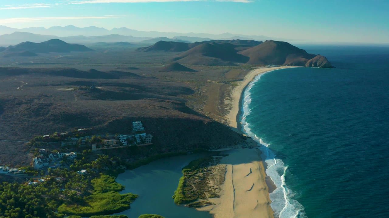 Wide drone view over Todos Santos, Baja California Sur, with the lagoon partly cut by the lower frame, buildings near trees, a long beach and surf line, dark coastal land, distant hills, and no visible people.