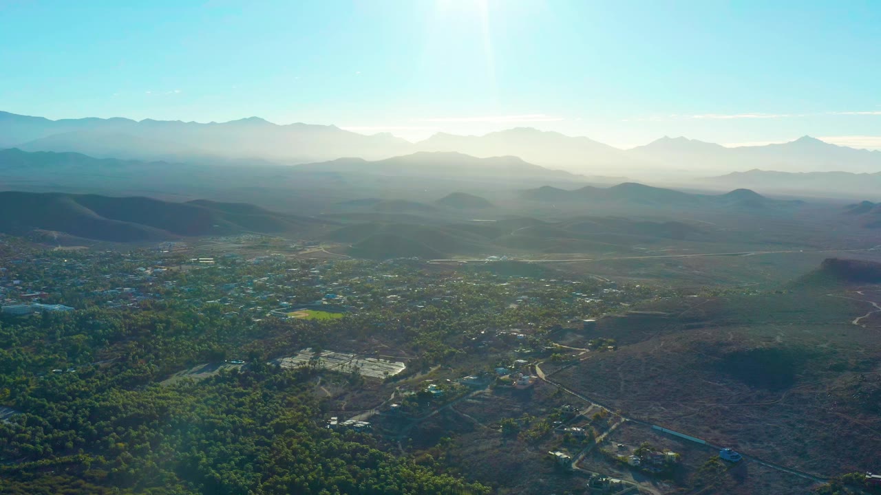 Wide aerial view over Todos Santos, Baja California Sur, with green tree cover, scattered buildings, dry hills, hazy mountain ridges, bright sun flare, and no visible people.