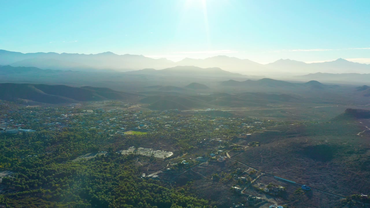 Wide aerial view over Todos Santos, Baja California Sur, with green foreground terrain, scattered town buildings, hazy mountain ridges, bright blue sky, and no visible people.
