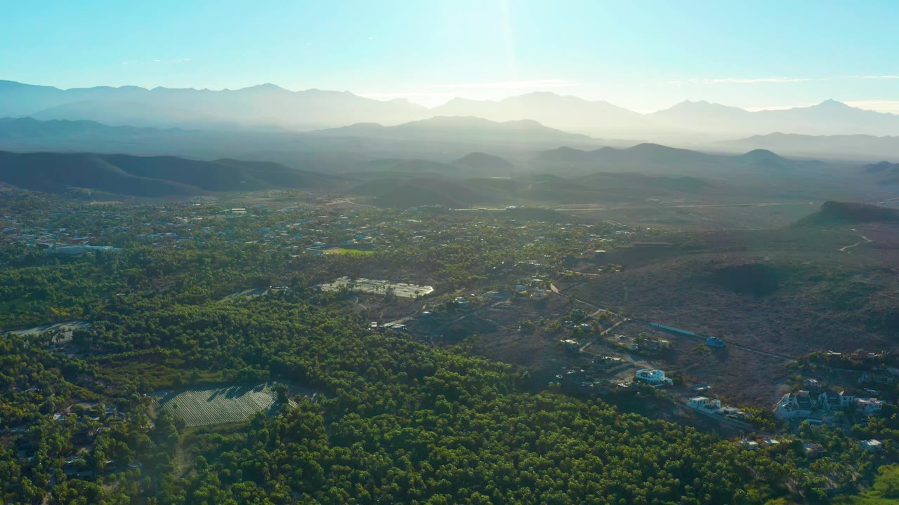 A high aerial frame over Todos Santos shows forested ground, brown open terrain, roads, scattered structures, hazy mountain ridges, and no visible people.