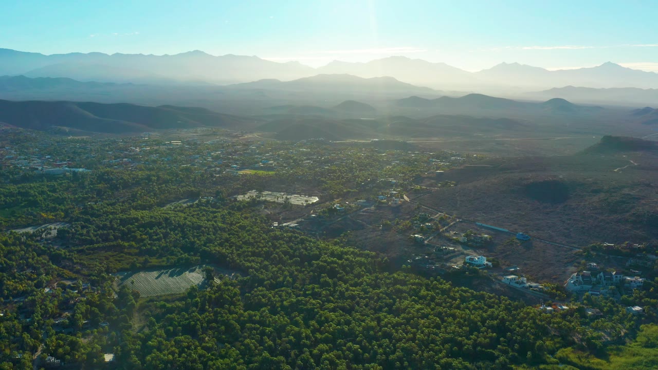 Sunlit Todos Santos aerial with dark green tree cover in the lower frame, pale open fields beyond, layered mountains in the background, and no visible people.