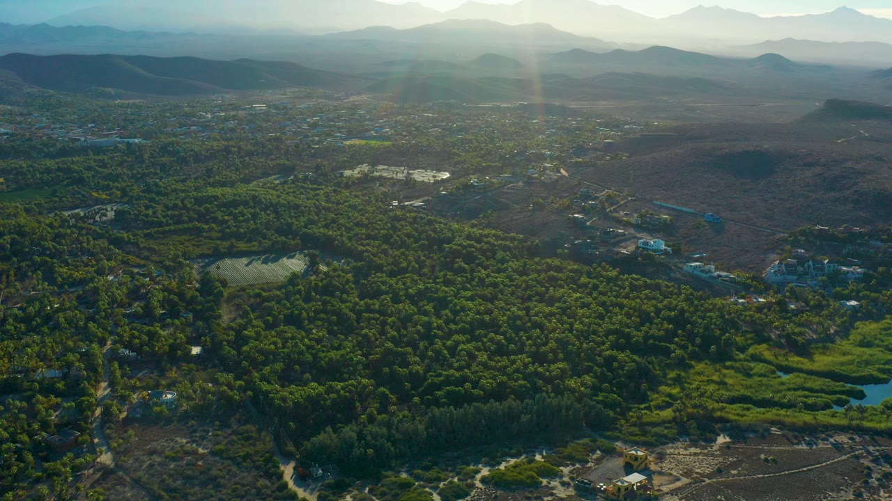 High aerial view over Todos Santos, Baja California Sur, with dense green forest, sandy beach at the bottom edge, brown open terrain, scattered buildings, hazy mountains, and no visible people.