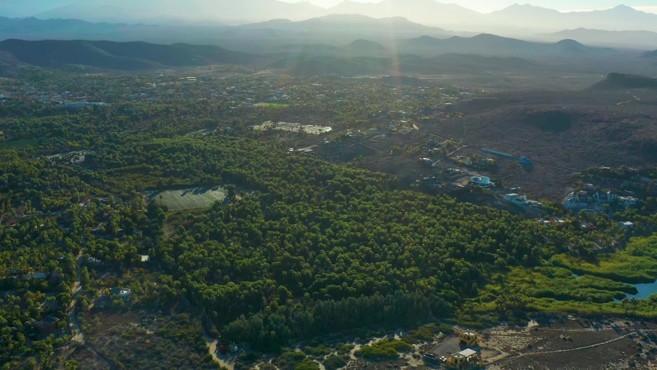 Aerial frame over Todos Santos showing the sandy coast along the lower edge, dense trees across the center, brown terrain to the right, scattered structures, distant mountain ridges, and no visible people.