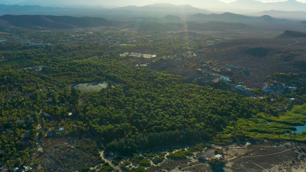Todos Santos aerial view with bright sun haze near the upper center, green forest and wetland-like patches in the middle, dry brown ground to one side, small buildings, mountains, and no visible people.
