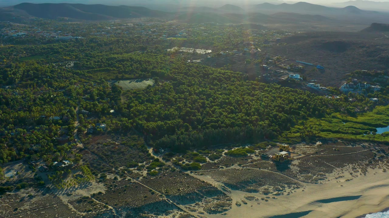 Aerial frame in Todos Santos showing a sandy beach edge, green forest, a small pond, dry scrub areas, distant hills, warm sun flare, and no visible people.