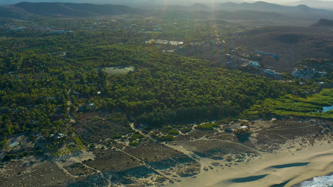 Aerial view over the Todos Santos coast with pale beach sand at the bottom, dense trees and a pond inland, scattered development, soft mountain silhouettes, and no visible people.