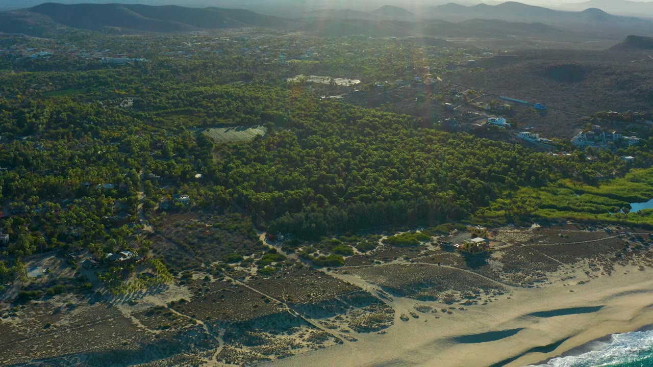 Mid-clip aerial frame over Todos Santos with more shoreline visible, including pale sand, a thin ocean edge, dense forest, a small pond, inland hills, and no visible people.