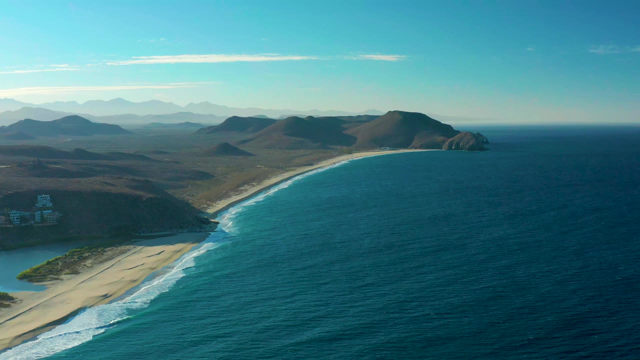 Opening aerial frame over Todos Santos, Baja California Sur, with a curved sandy beach, blue ocean, white surf lines, brown hills, distant mountains, and no visible people.