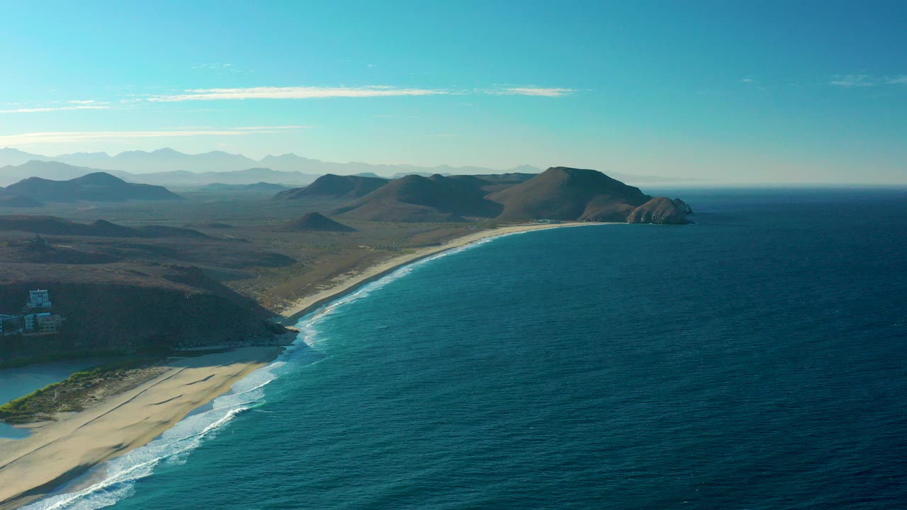 Aerial view over the Todos Santos coastline with a curved sandy beach, blue ocean to the right, dry hills inland, distant mountains, clear sky, and no visible people.