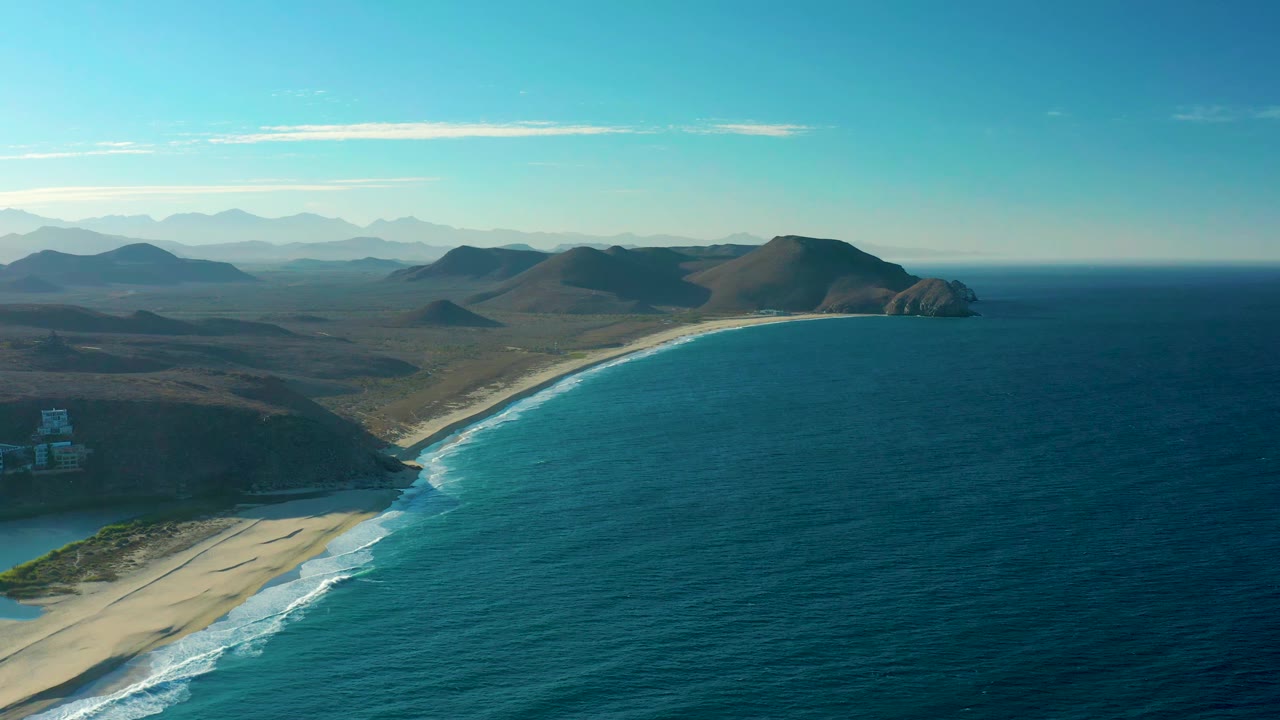 Wide drone view of surf breaking along a sandy shoreline beside hills.