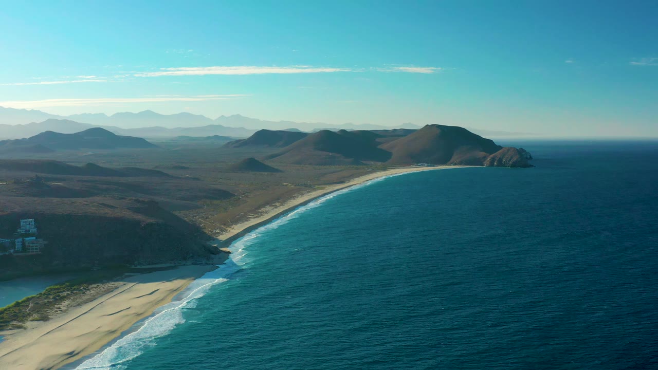 Aerial frame from Todos Santos showing white foam advancing along the sandy shore, deep blue water filling the right side, low hills in the midground, distant mountains, clear sky, and no visible people.