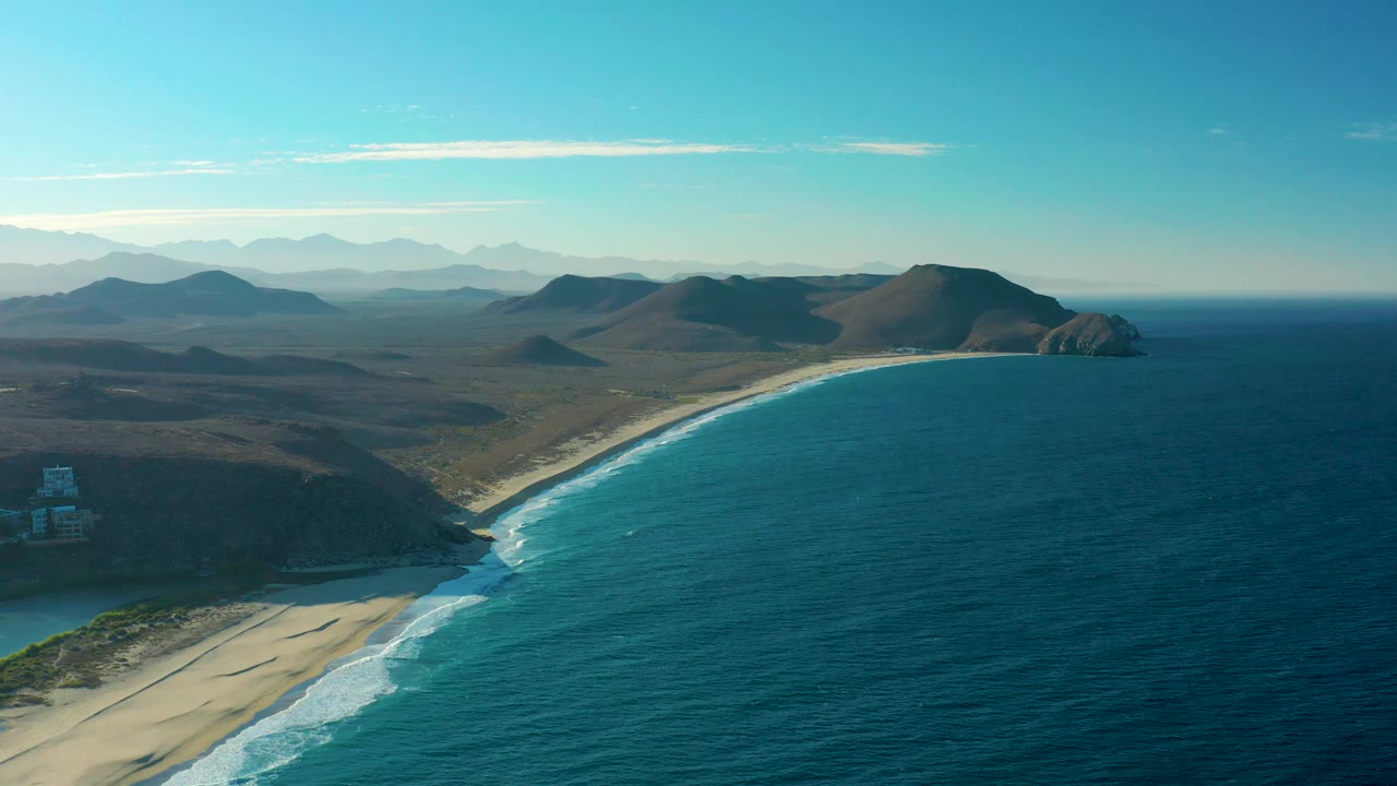 Drone-style aerial view over the Todos Santos shoreline with turquoise-blue water, a pale sandy beach, white wave edges, brown coastal hills, hazy mountain ridges in the background, and no visible people.