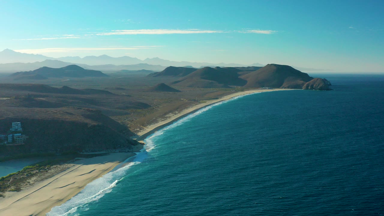 Opening aerial frame over Todos Santos with a broad blue bay, a pale sandy beach curving below, layered hills inland, and no visible people.