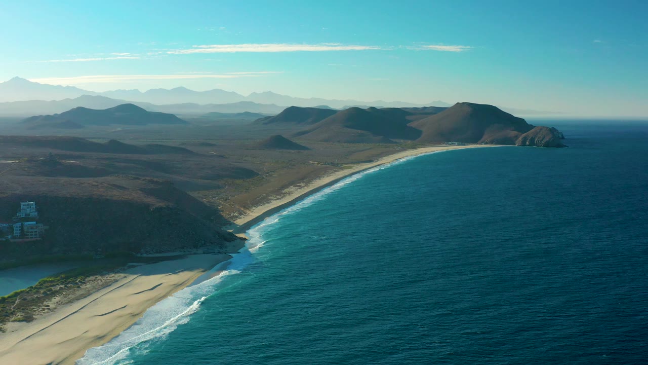 The frame holds on the Todos Santos bay with deep blue water filling the right side, a long arc of sand below the hills, and no visible people.
