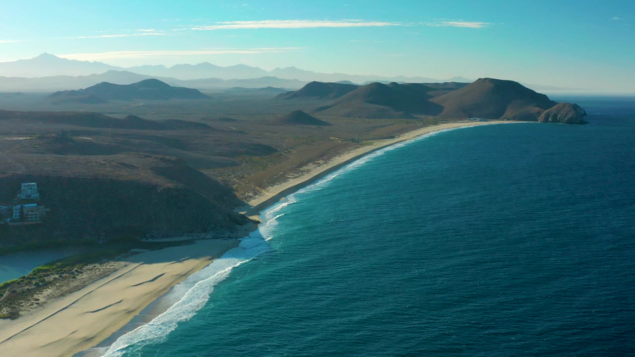 Aerial view over Todos Santos, Baja California Sur, showing a curved sandy beach, blue ocean, white surf, rugged hills, distant mountains, and no visible people.