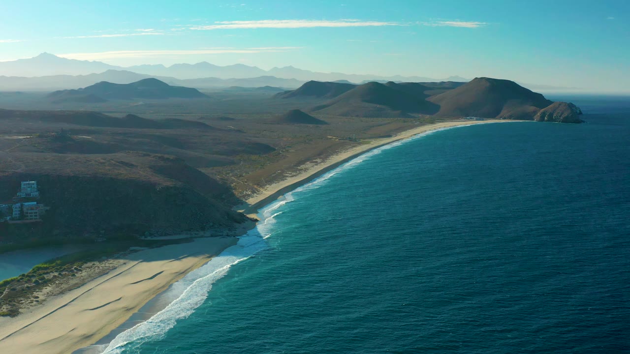 Wide aerial frame in Todos Santos with teal water on the right, pale sand below, scrub-covered hills inland, distant mountains on the horizon, and no visible people.