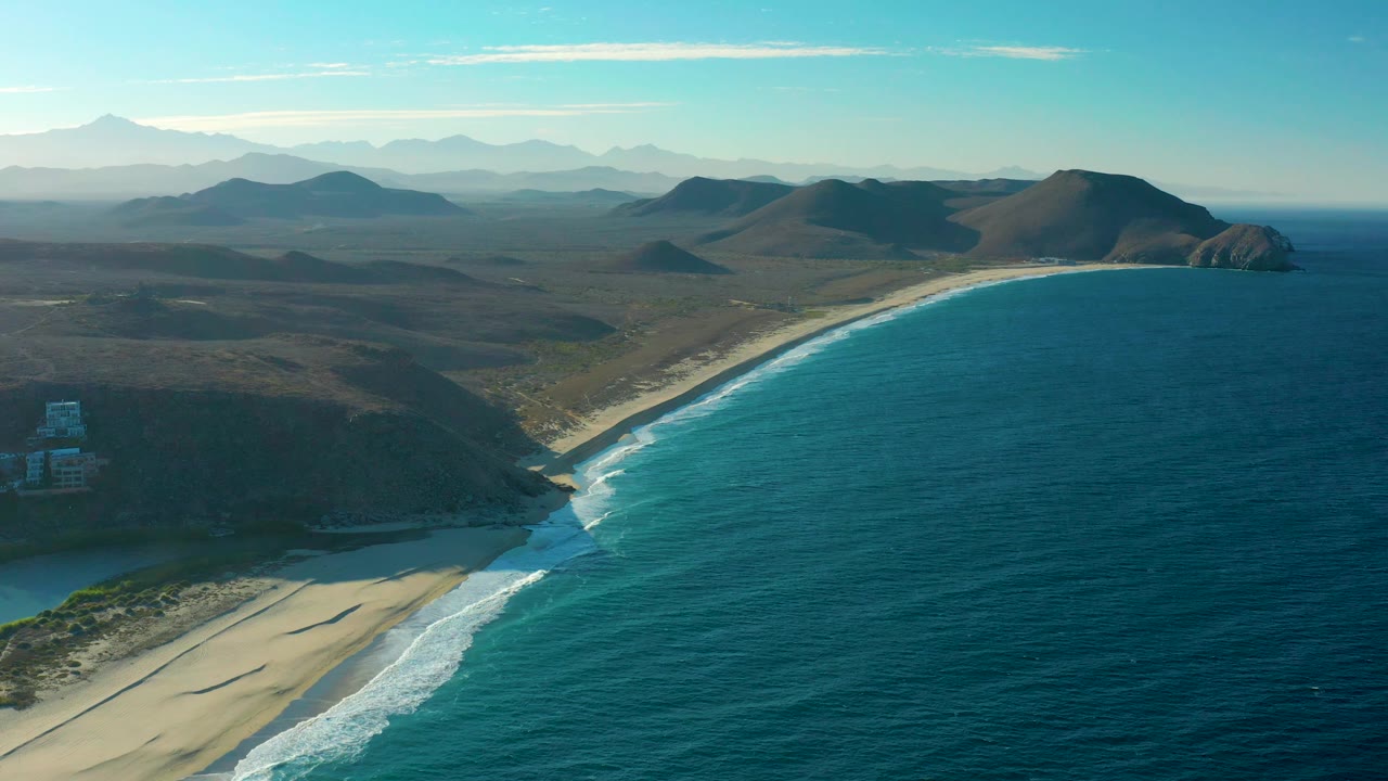 Elevated view of the Todos Santos shoreline with gentle waves along the sand, dry inland hills, layered mountains in the distance, bright blue sky, and no visible people.