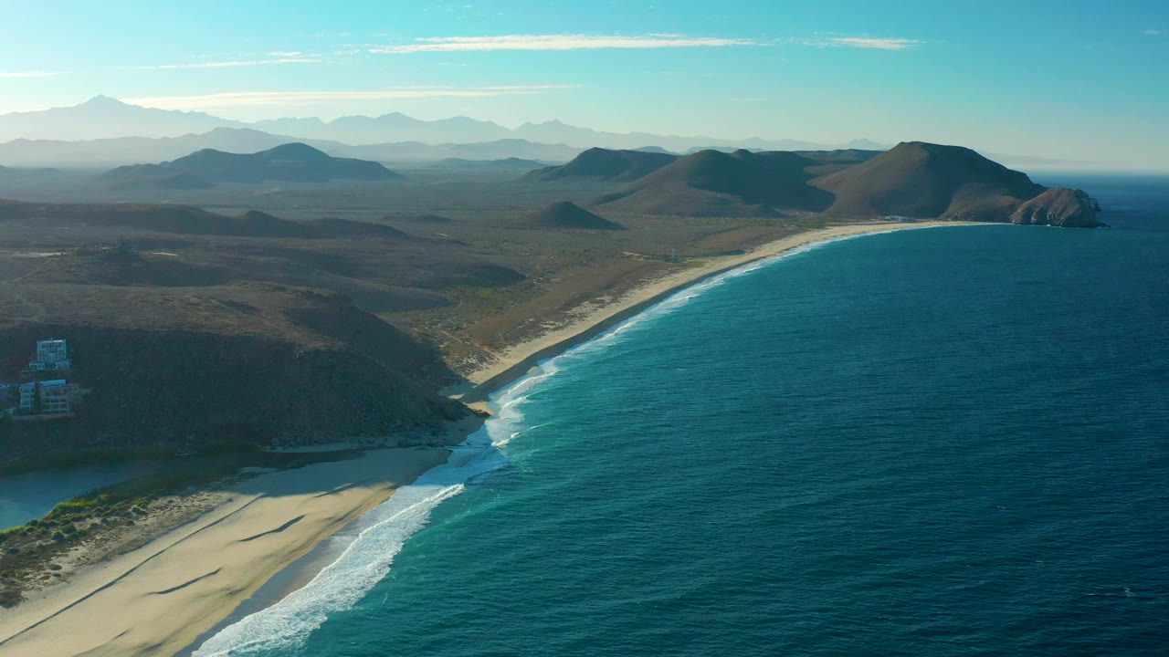 Aerial view in Todos Santos with the ocean filling the right half, sandy beach at the bottom, darker coastal hills to the left, distant mountains beyond, and no visible people.