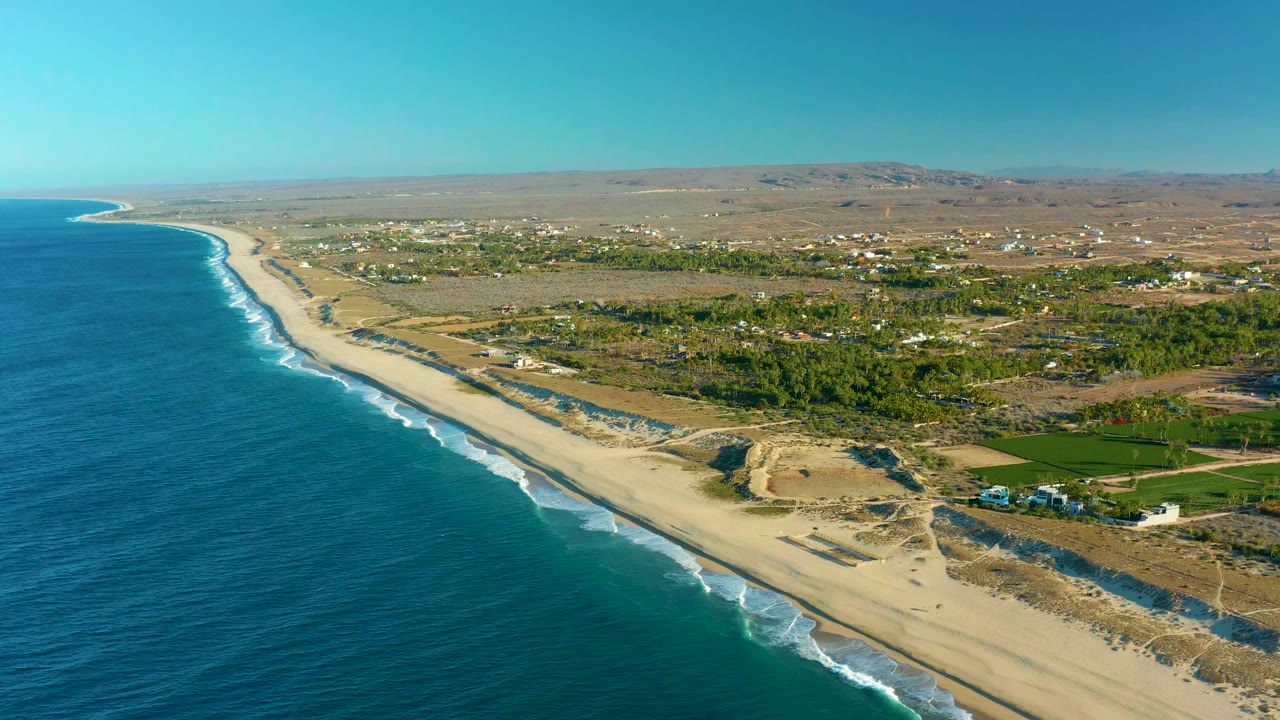 Aerial view of the Todos Santos shoreline one second into the clip, with parallel wave lines along the sand, a green inland strip, scattered white buildings, dry hills, and no visible people.
