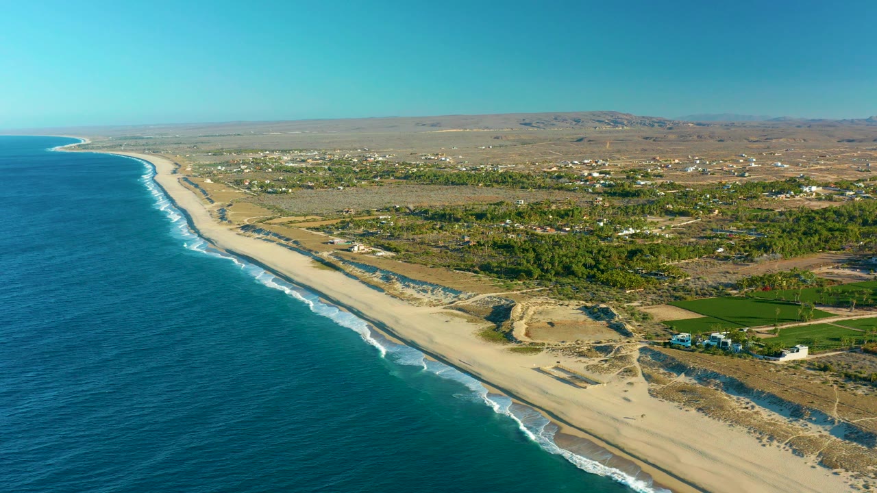 Drone view of sandy beach, ocean surf, dunes, fields, and buildings.