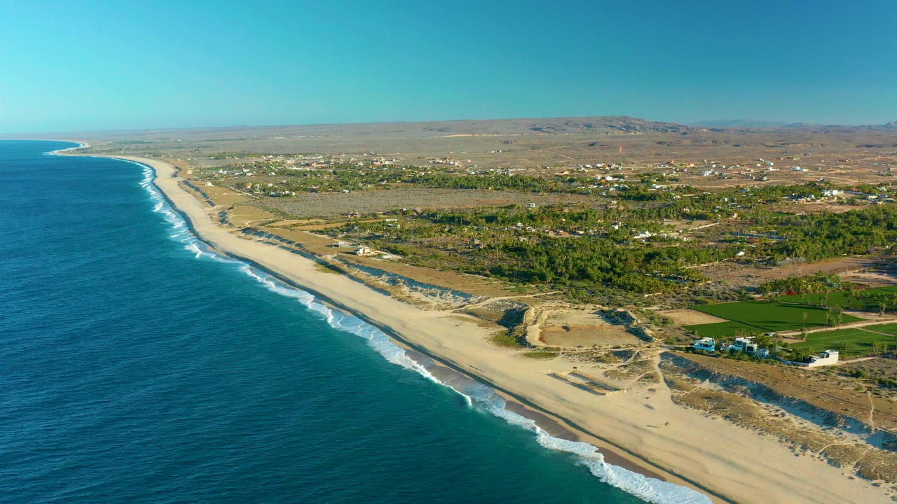 Aerial frame over the Todos Santos, Baja California Sur coast with blue ocean on the left, white surf reaching a long sandy beach, inland vegetation, scattered buildings, and no visible people.