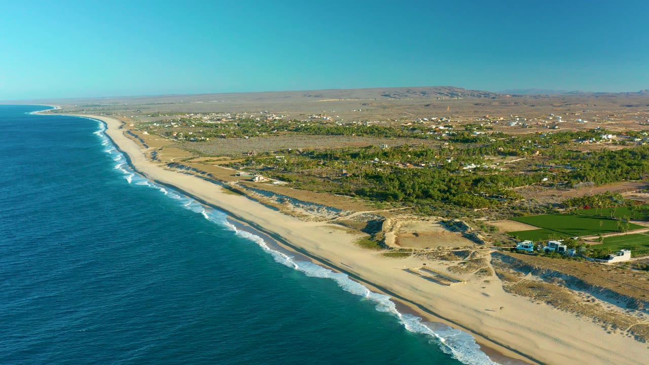Opening aerial frame over Todos Santos, Baja California Sur, with blue ocean on the left, white surf touching a long sandy beach, inland vegetation and scattered buildings beyond, and no visible people.