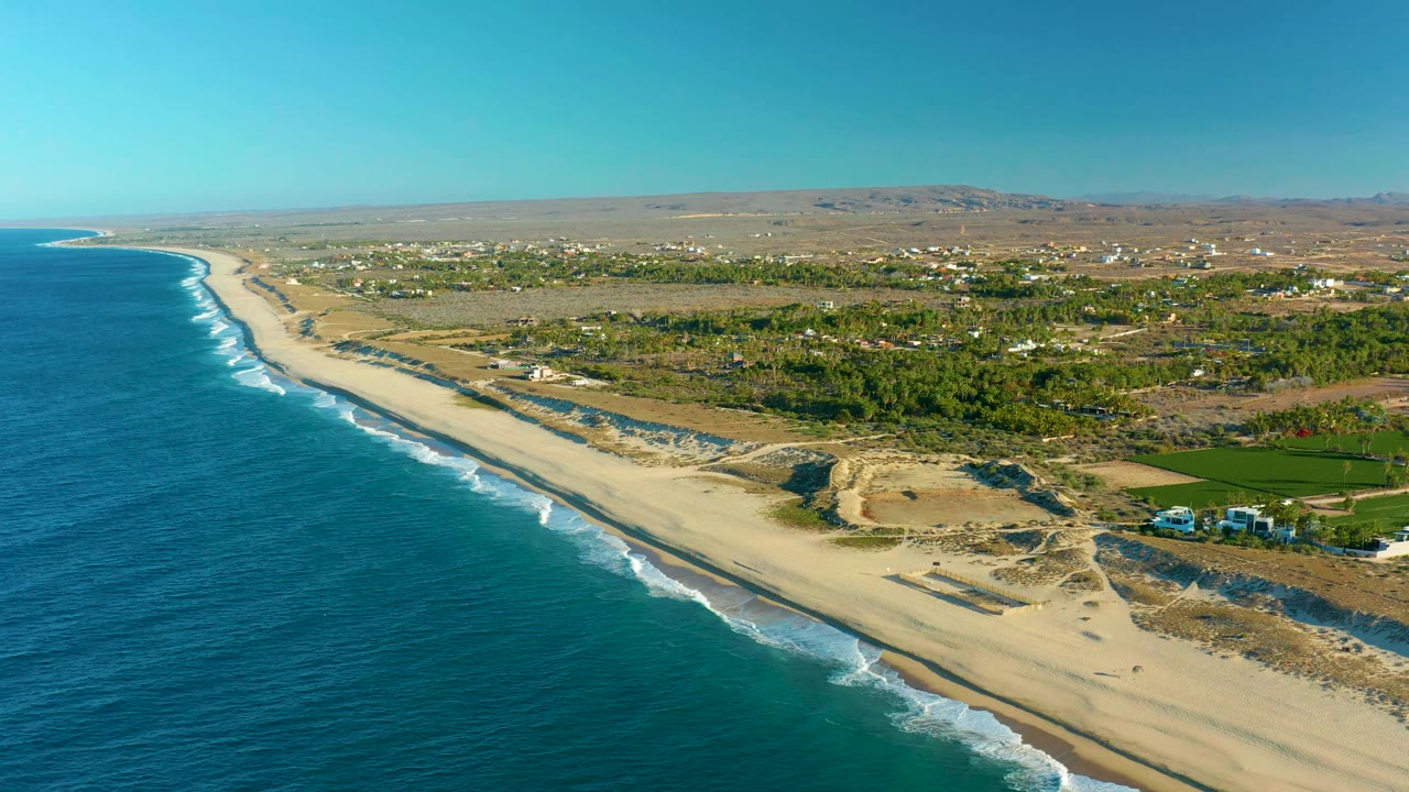 Wide drone view of ocean surf beside a long sandy beach and inland greenery.