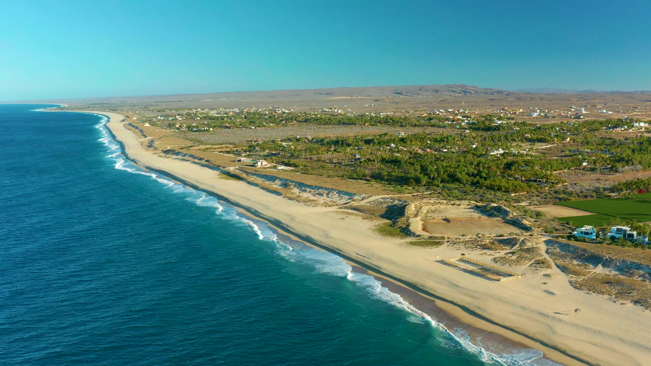 Aerial frame over Todos Santos coastline showing the ocean filling the left side, parallel wave breaks along the sand, dune tracks near the beach, green inland vegetation, and no visible people.