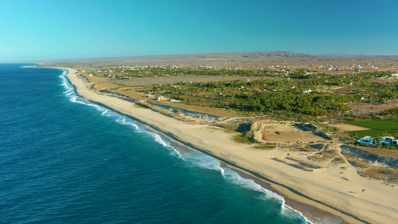 High aerial view of ocean waves meeting a long sandy beach with dunes and inland greenery.