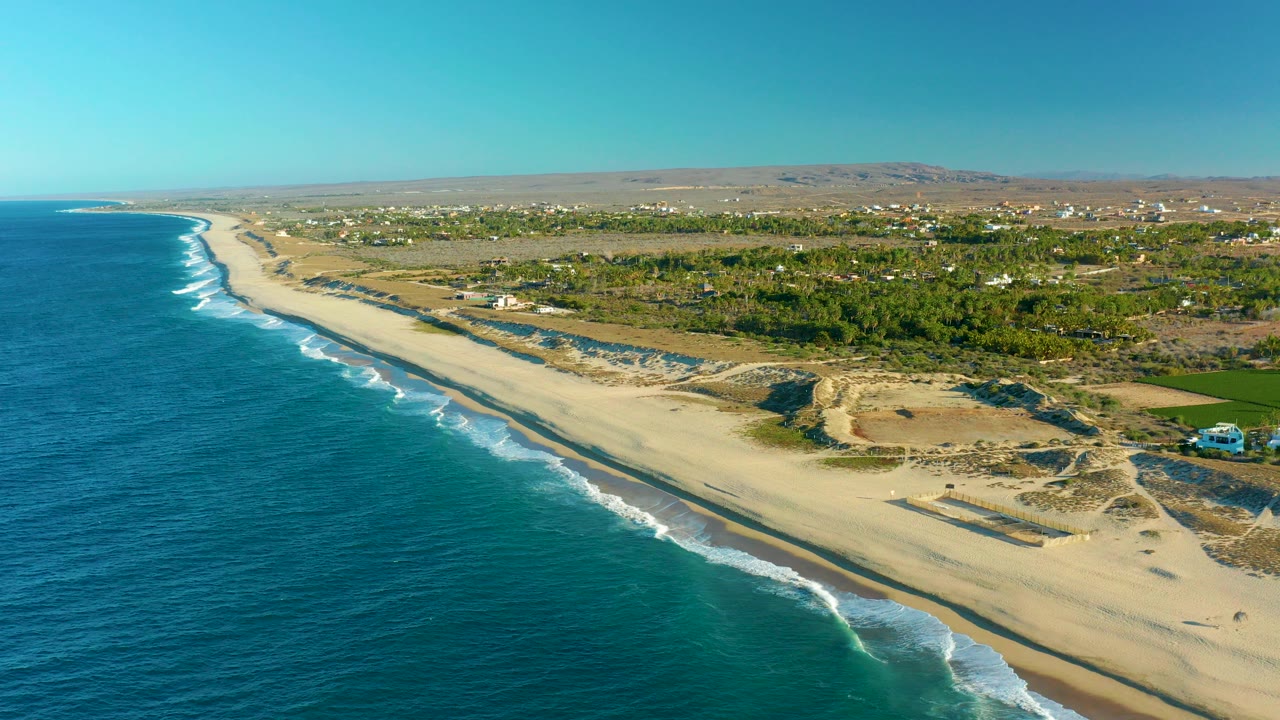 Aerial view over Todos Santos, Baja California Sur, with blue ocean on the left, white surf approaching a sandy beach, greenery and scattered buildings inland, and no visible people.
