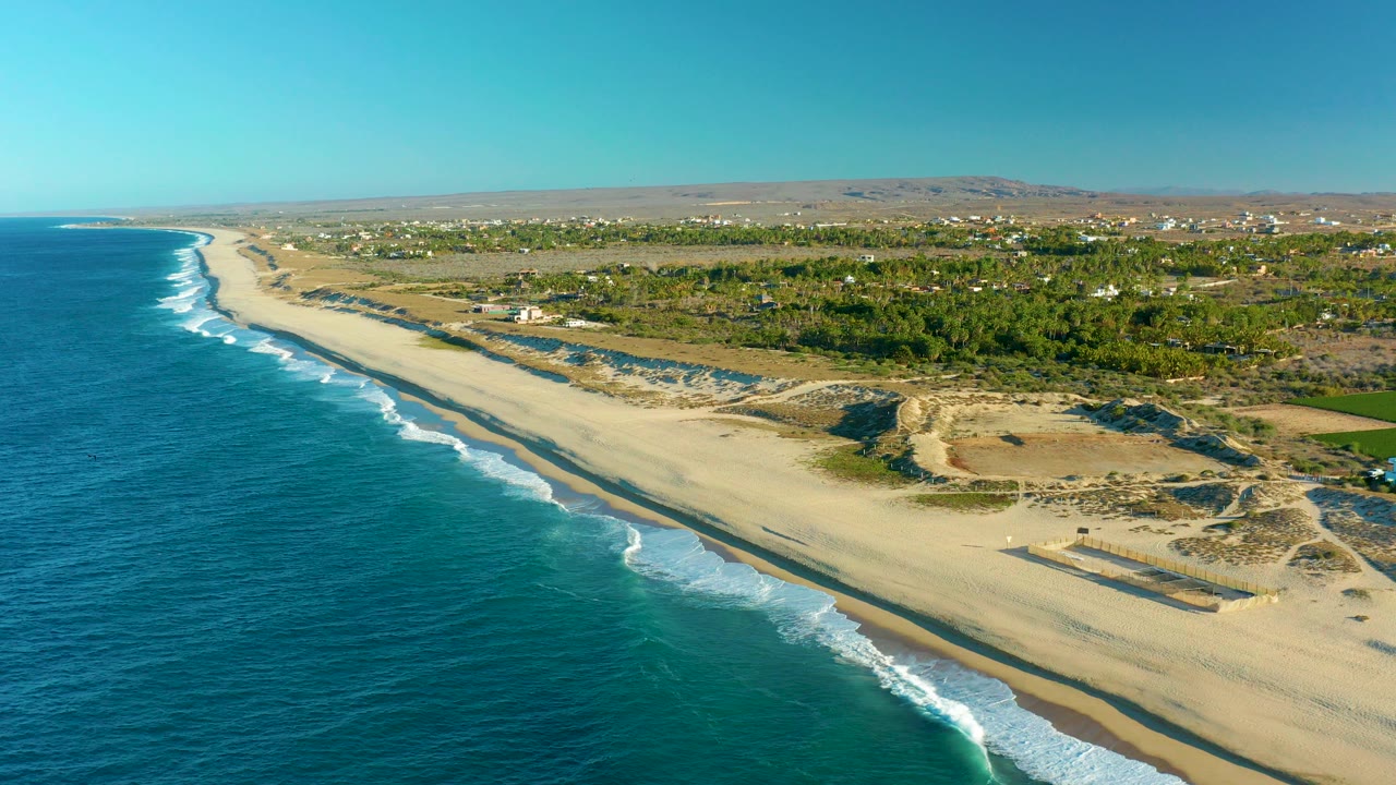 Aerial frame over Todos Santos, Baja California Sur, Mexico, showing blue ocean at left, white surf lines on a sandy beach, dune tracks, green vegetation, distant buildings, and no visible people.