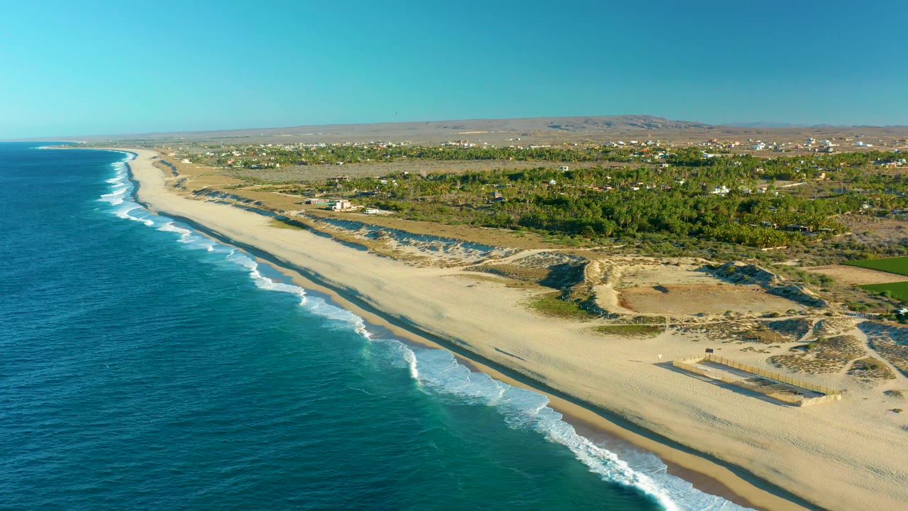 Aerial coastal frame from Todos Santos showing blue ocean, white wave breaks, a wide pale beach, dune paths, inland green vegetation, distant buildings, dry hills, and no visible people.