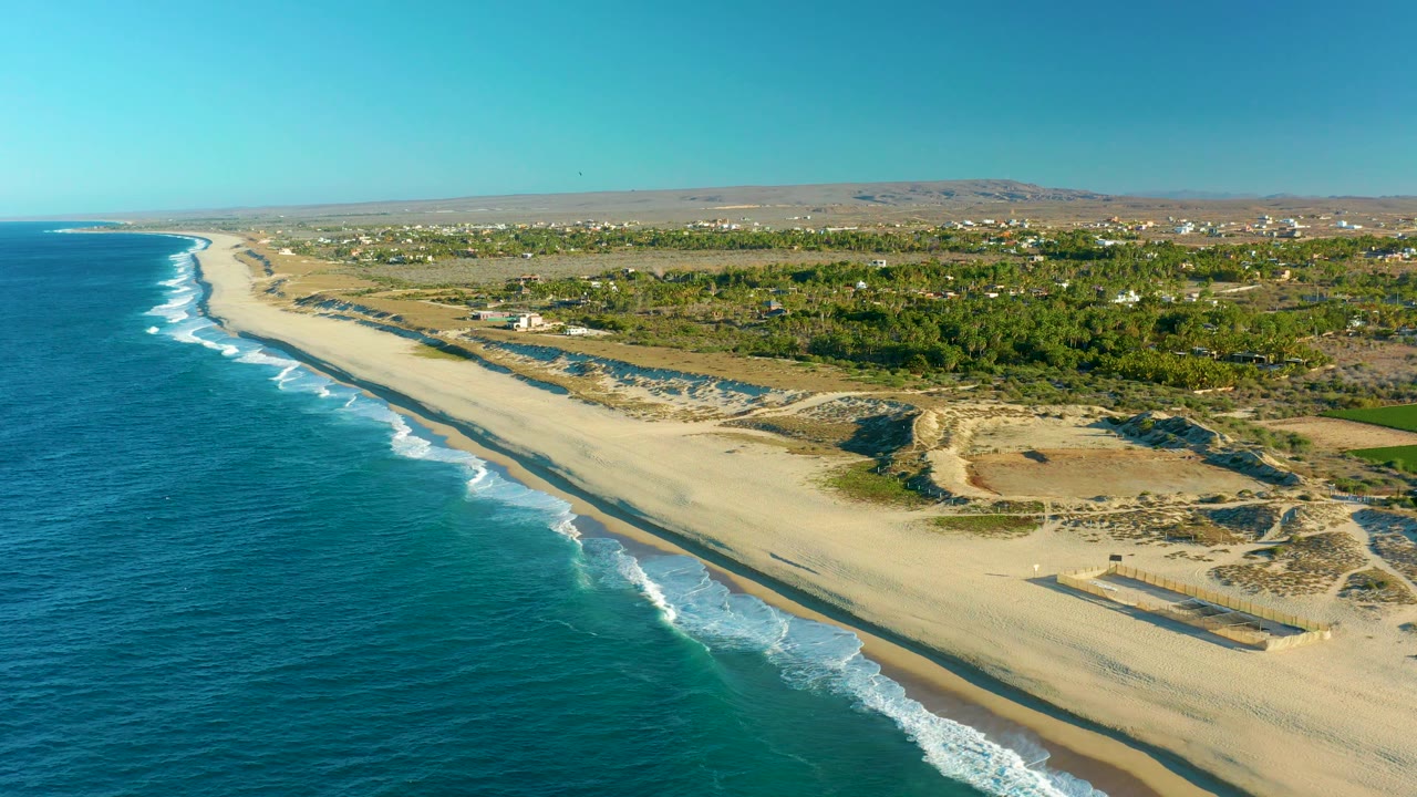 Aerial coastline with white surf, sandy beach, dune area, vegetation, and distant homes.
