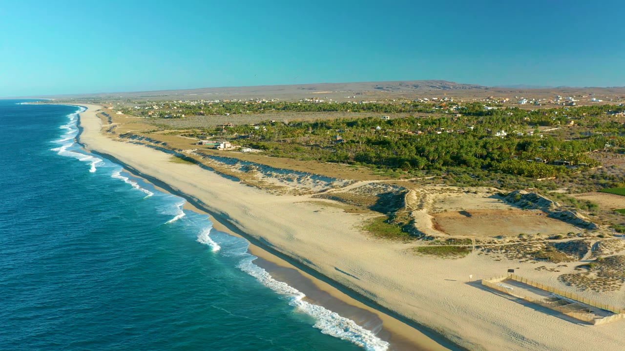 Aerial view along the Todos Santos shoreline, with surf bands moving toward pale sand, dune tracks and vegetation inland, distant hills, and no visible people.
