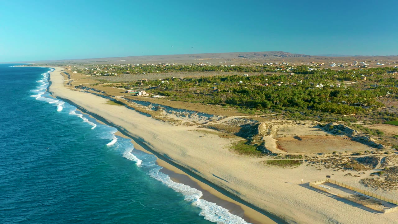 A high coastal frame from Todos Santos shows turquoise water and breaking waves beside a broad beach, low dunes, scattered inland buildings, dry hills, and no visible people.