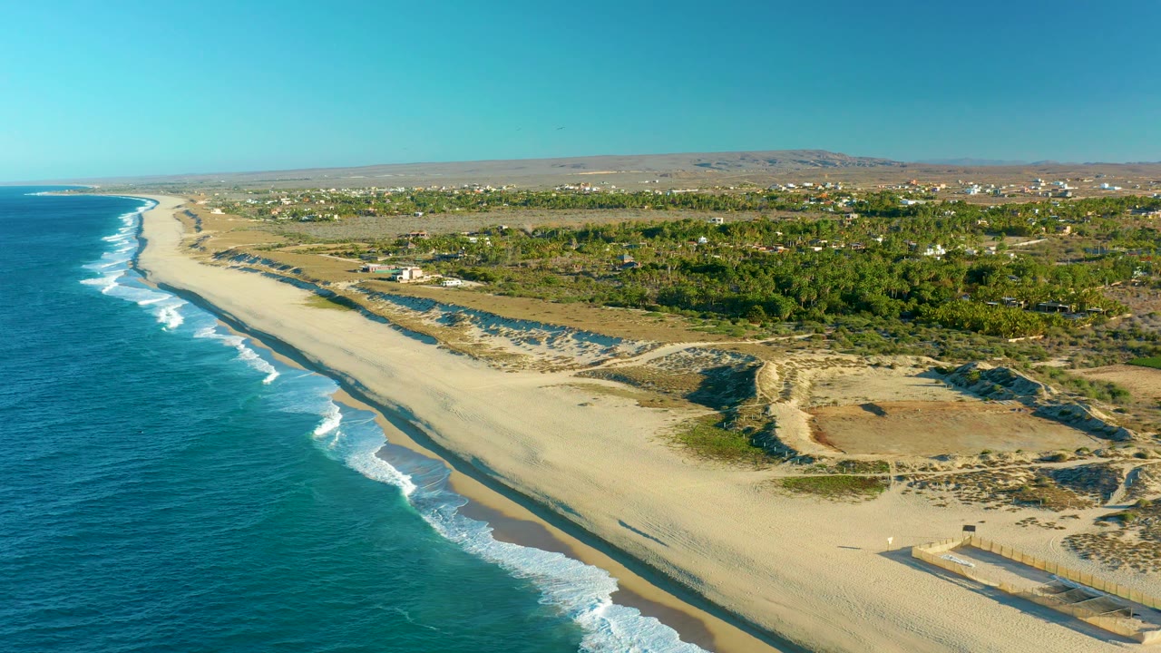 Mid-clip aerial view over Todos Santos coastline, with the ocean filling the left side, foamy waves edging the sand, inland green vegetation and dunes to the right, and no visible people.