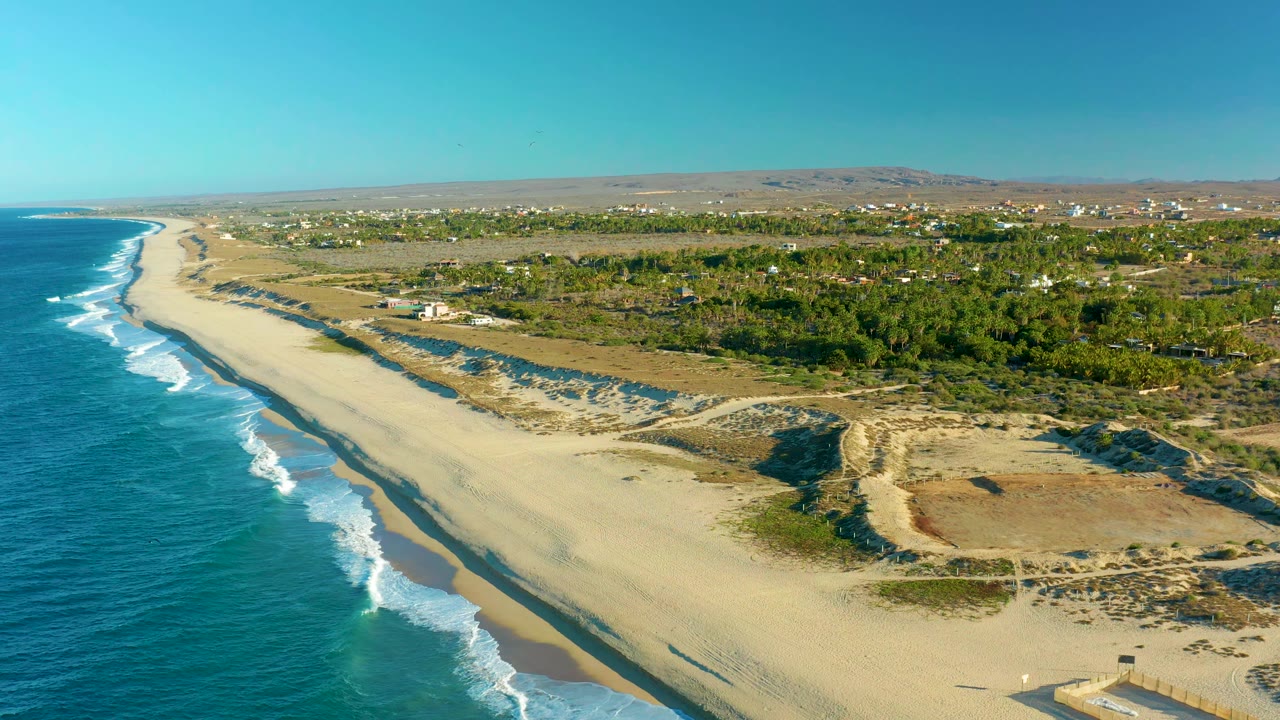 Aerial view in Todos Santos, Baja California Sur, with the ocean running along the left edge, surf breaking diagonally onto pale sand, dune paths in the lower right, inland greenery, distant structures, open blue sky, and no visible people.