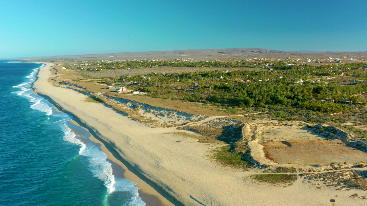 Aerial frame from Todos Santos, Baja California Sur, where the surf line sits farther down the left edge, the wide beach fills the center, dunes and scrub appear in the lower right, inland vegetation and distant structures sit under clear blue sky, and no visible people appear.