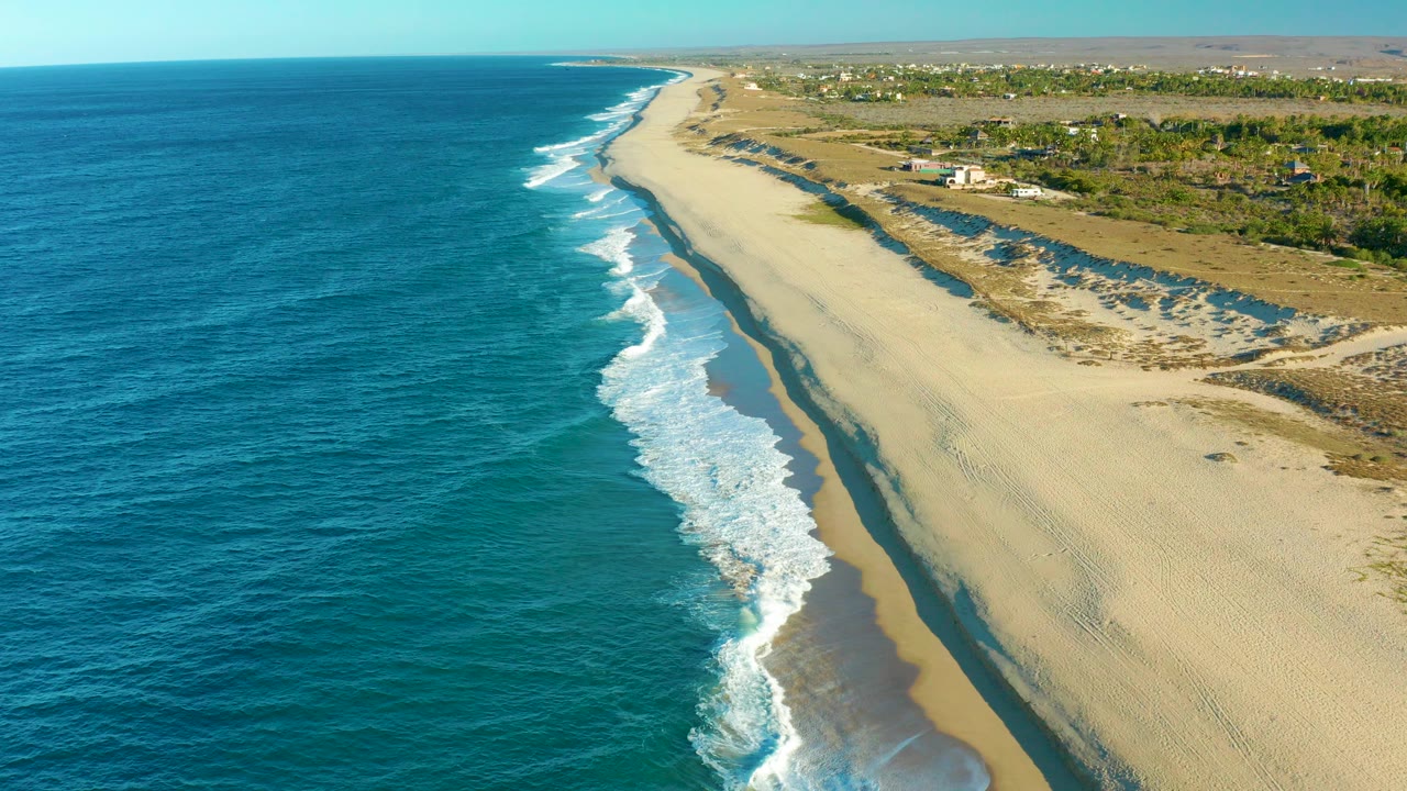 Aerial frame over Todos Santos, Baja California Sur, with blue ocean on the left, white surf reaching a broad sand beach, dunes inland, scattered buildings near the horizon, and no visible people.