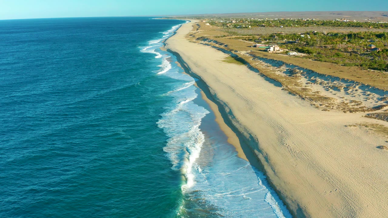 Opening frame over the Todos Santos coastline, with blue ocean on the left, white surf reaching the sand, dunes and distant structures inland, and no visible people.
