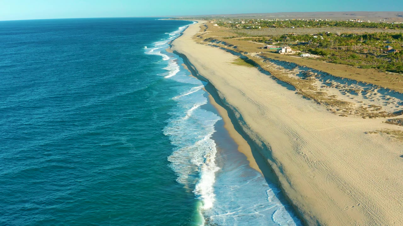 Todos Santos beach appears from above with deeper blue water at left, a fresh line of white foam near center, open sand at right, inland scrub, and no visible people.