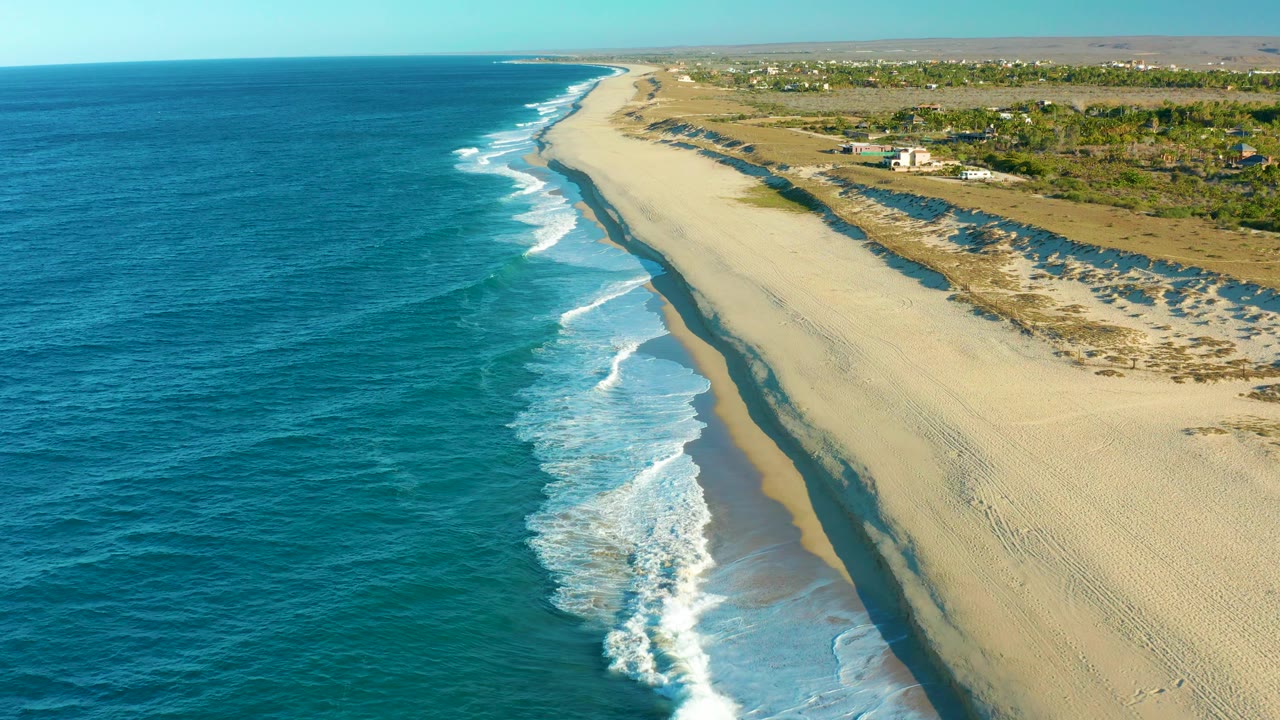 Aerial coastline view with surf, pale sand, dunes, and distant buildings.
