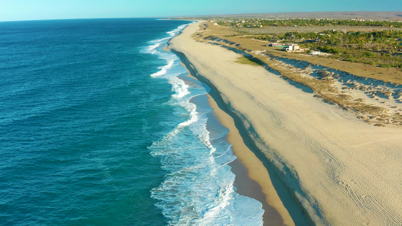 Aerial view in Todos Santos showing a long line of foam advancing onto the beach, with blue water filling the left half and low coastal vegetation beyond the sand, no visible people.
