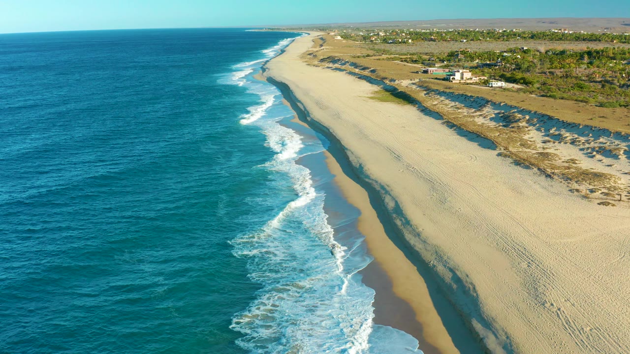 The Todos Santos coastline is seen from above with white wave foam near the center, pale sand to the right, dry dunes and a few small distant structures inland, and no visible people.