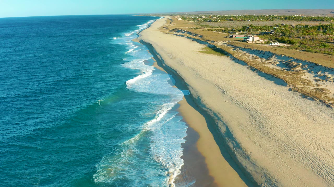At 0:00, an aerial view of the Todos Santos shoreline shows blue ocean on the left, white surf reaching a broad sandy beach, sparse coastal vegetation, distant structures, and no visible people.