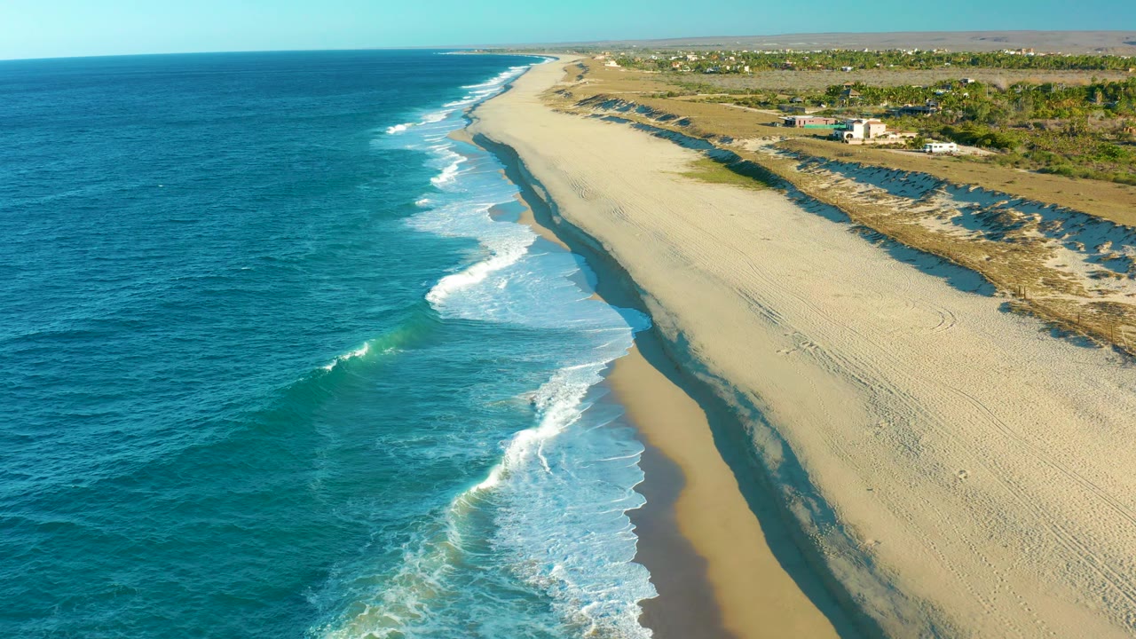 At 0:02, the aerial view looks along the Todos Santos coast as foam spreads in curved bands along the shore, with a long empty sand strip and low inland vegetation visible, no visible people.