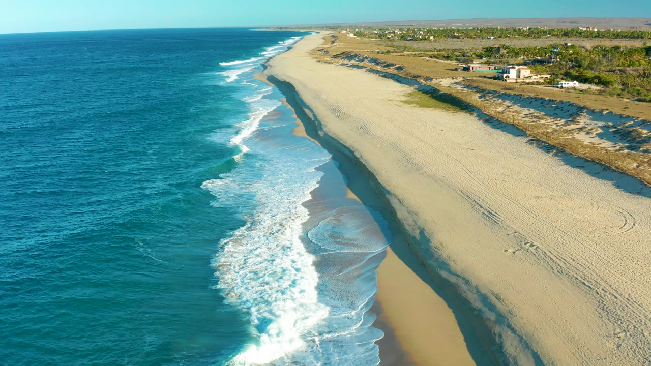 At 0:00 in Todos Santos, Baja California Sur, blue ocean fills the left side while white surf reaches the sand and tire tracks cross the beach, with no visible people.