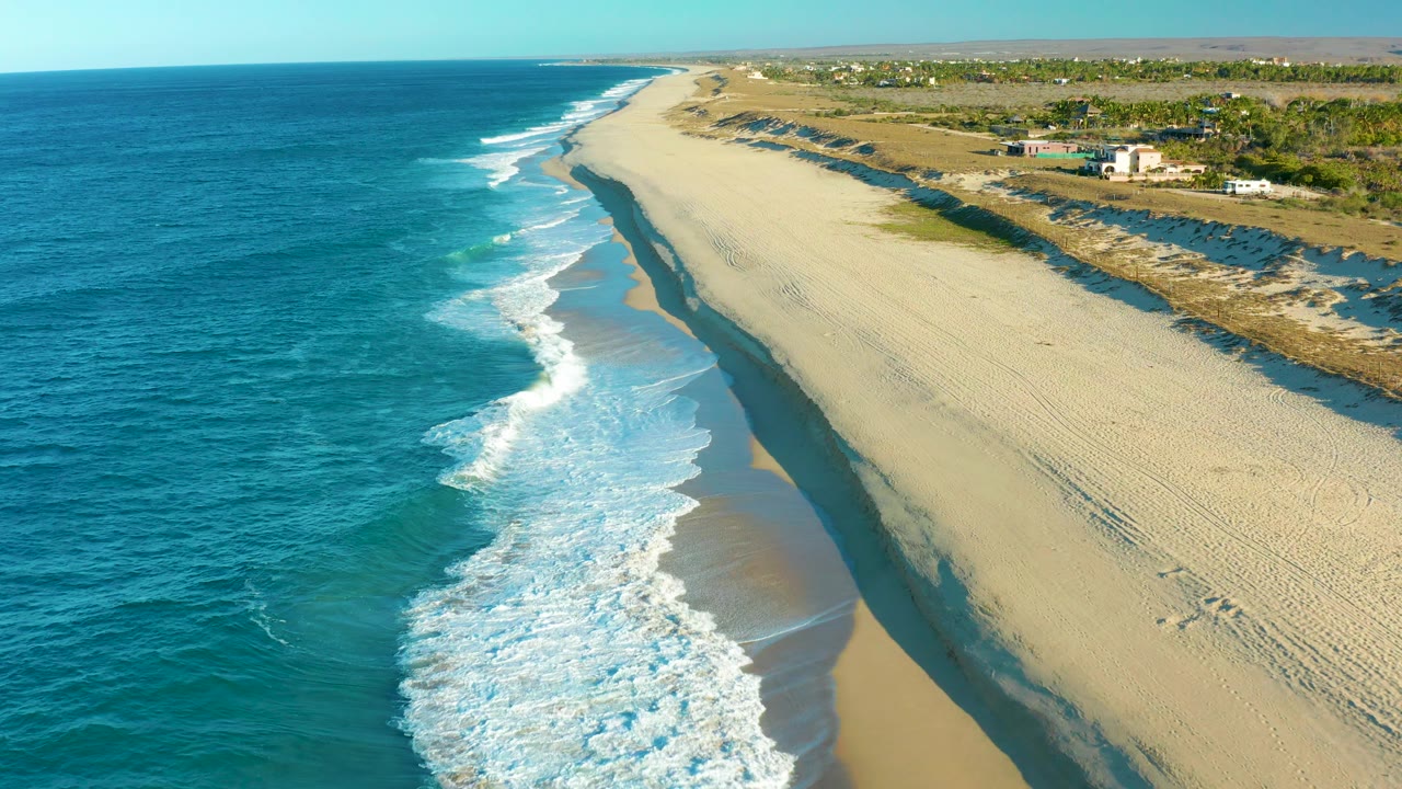 Aerial coastline view with waves, empty sand, and tire tracks near dunes.