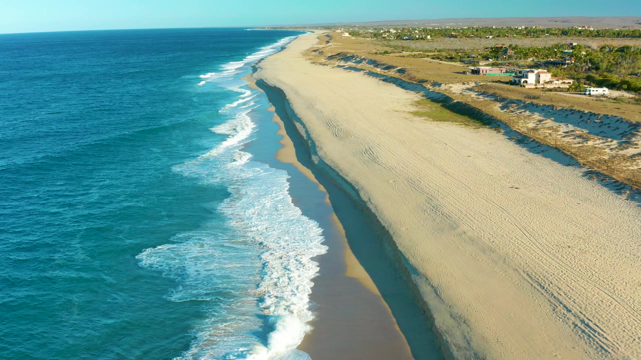 Aerial view over Todos Santos, Baja California Sur, with turquoise ocean on the left, white surf meeting a wide sandy beach, inland vegetation and scattered buildings beyond, and no visible people.